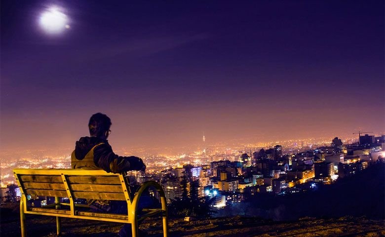 تهران در شب از زاویه دید مرد جوان روی نیمکتی در بام تهران