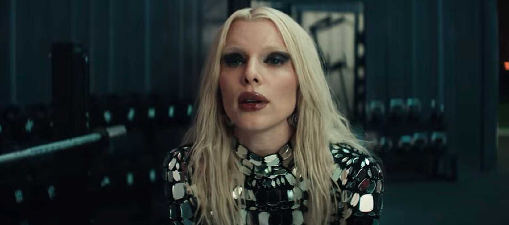 جولیا فاکس در فیلم Him، در لباسی از پنل‌های شیشه‌ای گرد، با موهای بلوند، رژ لب قرمز تیره و سایه چشم تیره